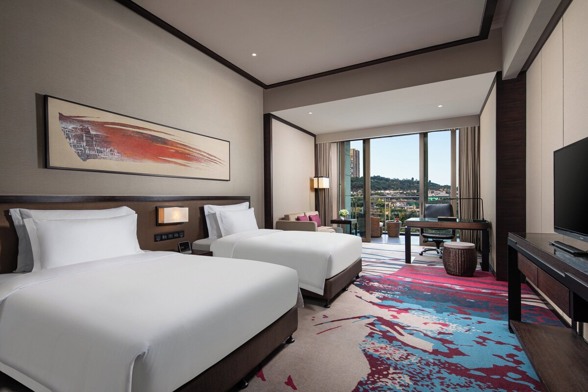 Habitación de hotel con dos camas, un escritorio, un televisor y un ventanal con vistas a la ciudad.