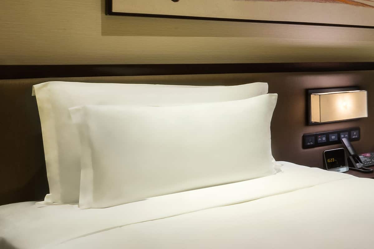 A hotel room with a neatly made bed, a bedside table with a lamp, and a digital clock displaying the time.