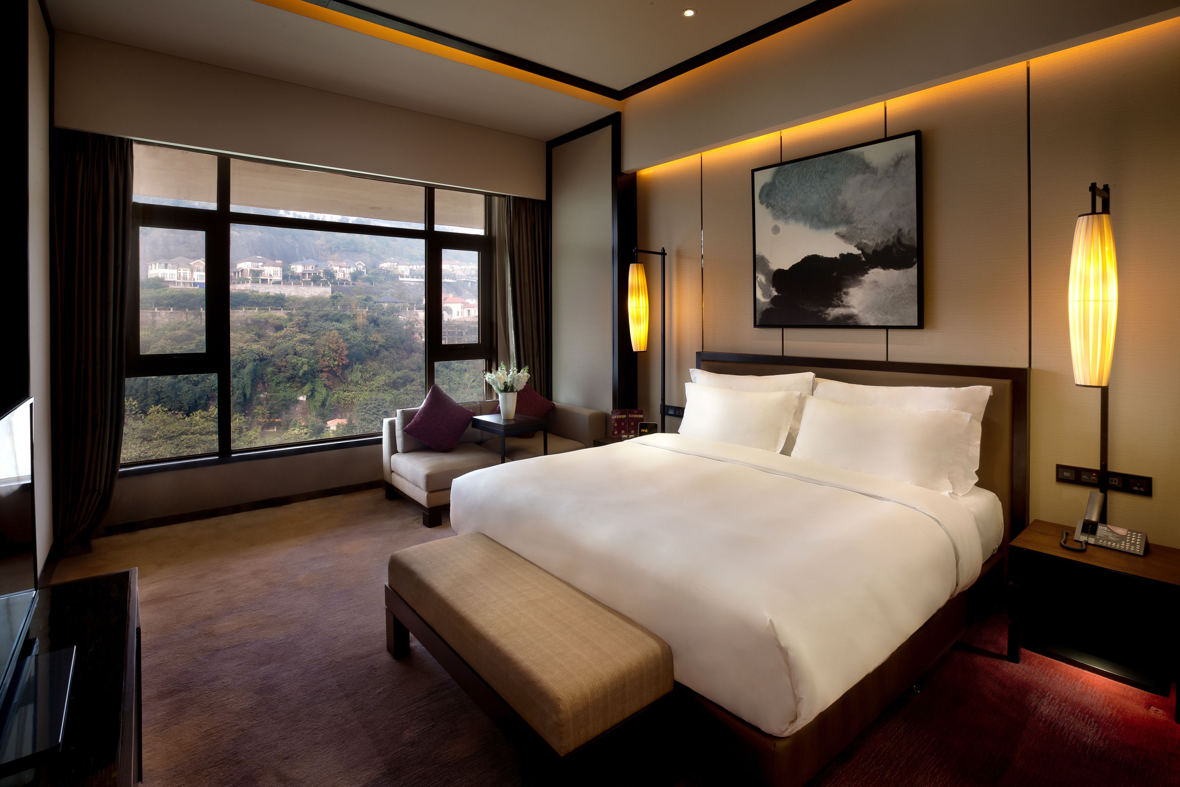 A hotel room with a large bed, a bench, a desk, and a view of a cityscape through large windows.