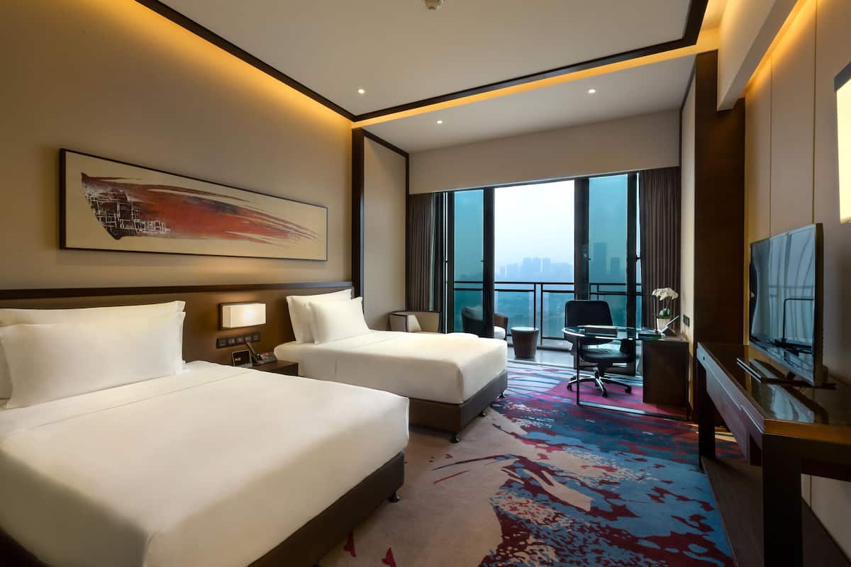 A hotel room with two beds, a desk, a chair, a TV, and a large window with a city view.