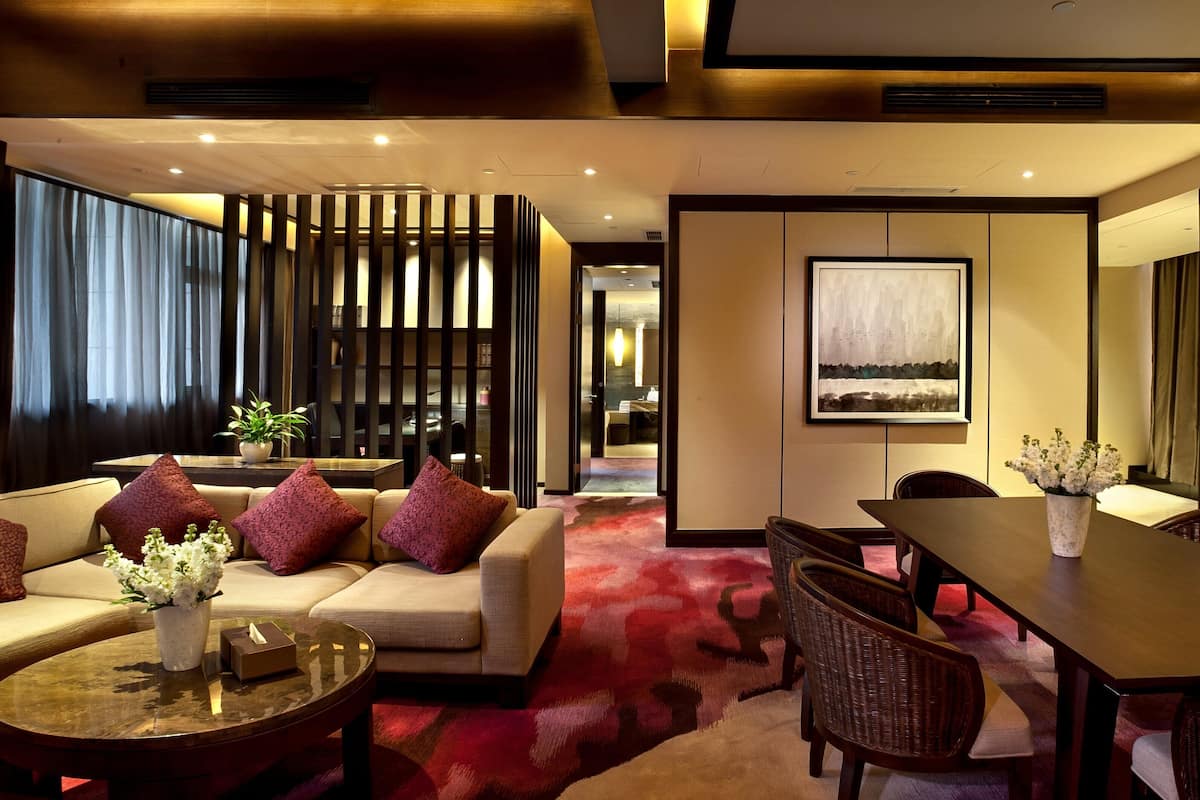 A modern hotel room with a sofa, dining table, and chairs. There is a large window with curtains, a painting on the wall, and a potted plant.