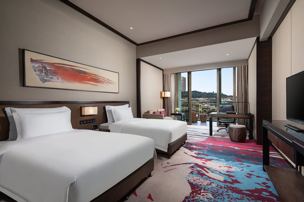 A hotel room with two beds, a desk, a TV, and a large window offering a city view.