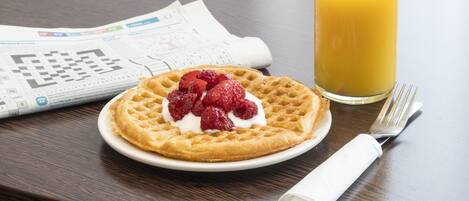 A waffle with strawberries and whipped cream on a plate, a glass of orange juice, and newspapers on a table.