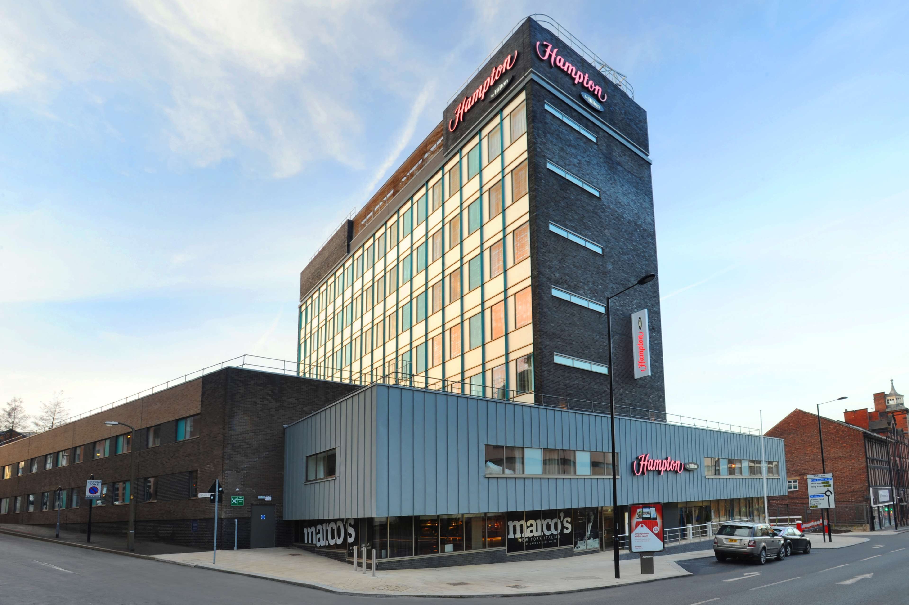 A Hampton hotel with a modern facade, multiple windows, and a visible storefront with a sign for 'marco's'.