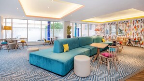 A modern hotel lobby with a sectional sofa, round tables, and chairs, large windows, and a geometric-patterned wall. - Hampton By Hilton Sheffield (Sheffield)