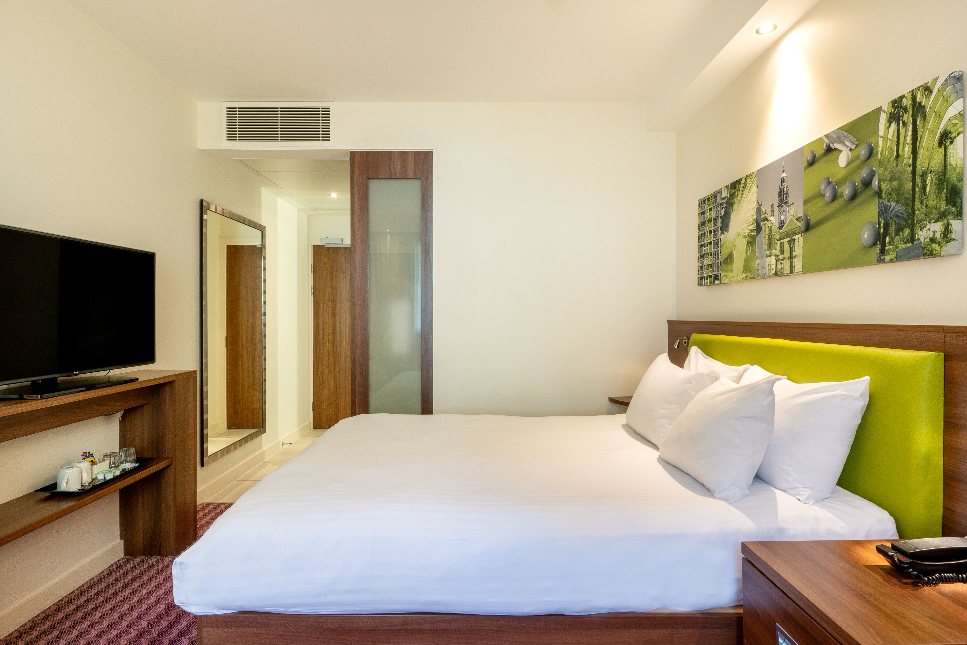 A hotel room with a large bed, a flat-screen TV, a wooden nightstand, a mirror, and a painting on the wall.
