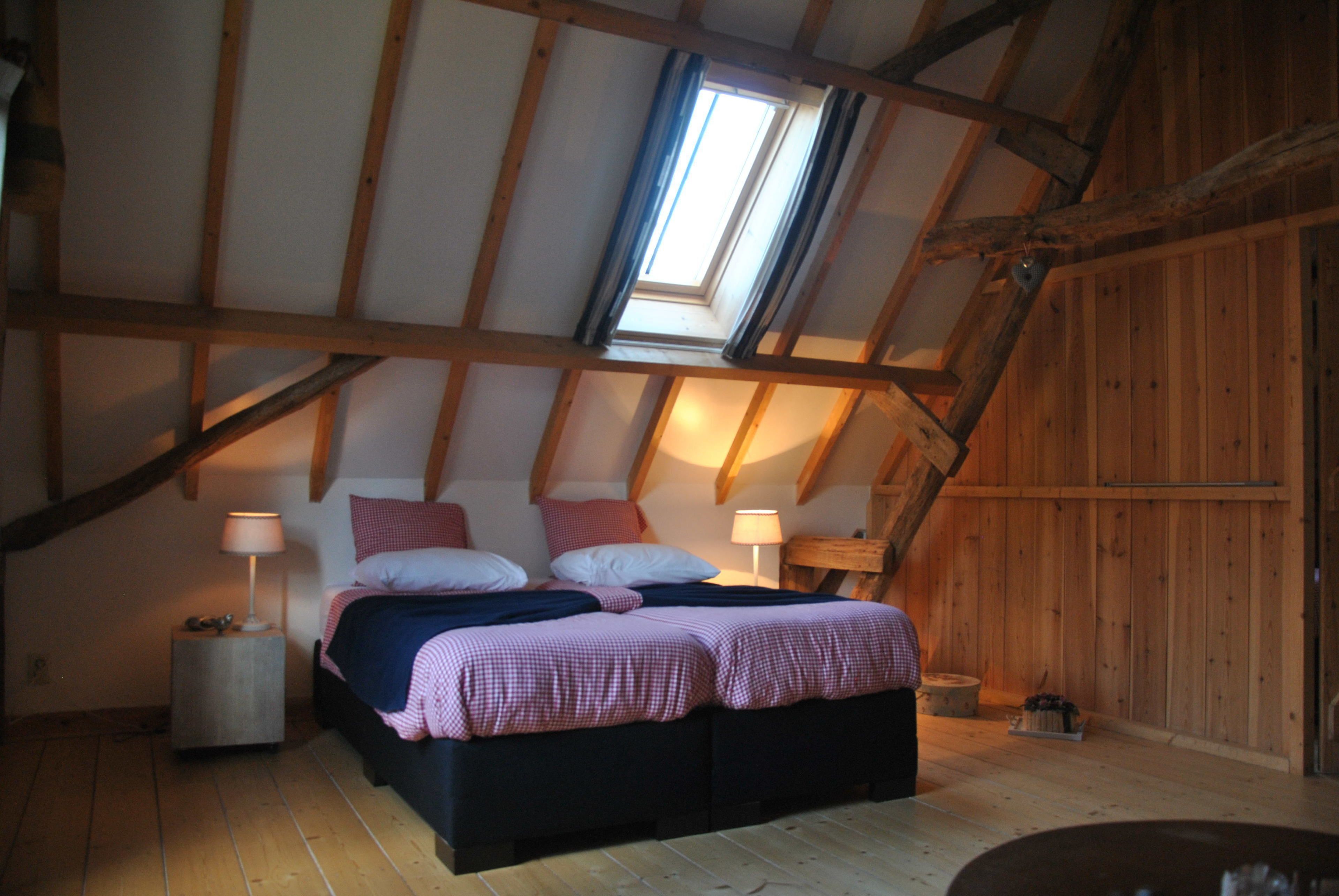 Un dormitorio con techo inclinado, tragaluz, cama con ropa de cama rosa y blanca, dos mesitas de noche con lámparas y paredes de madera.