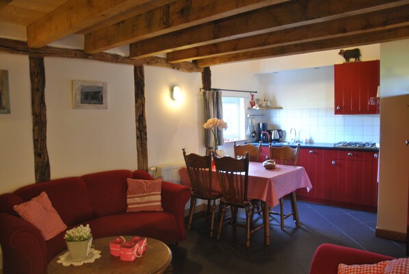 A cozy living room with a red sofa, wooden dining table, and chairs. A kitchen with red cabinets and a window is visible in the background.
