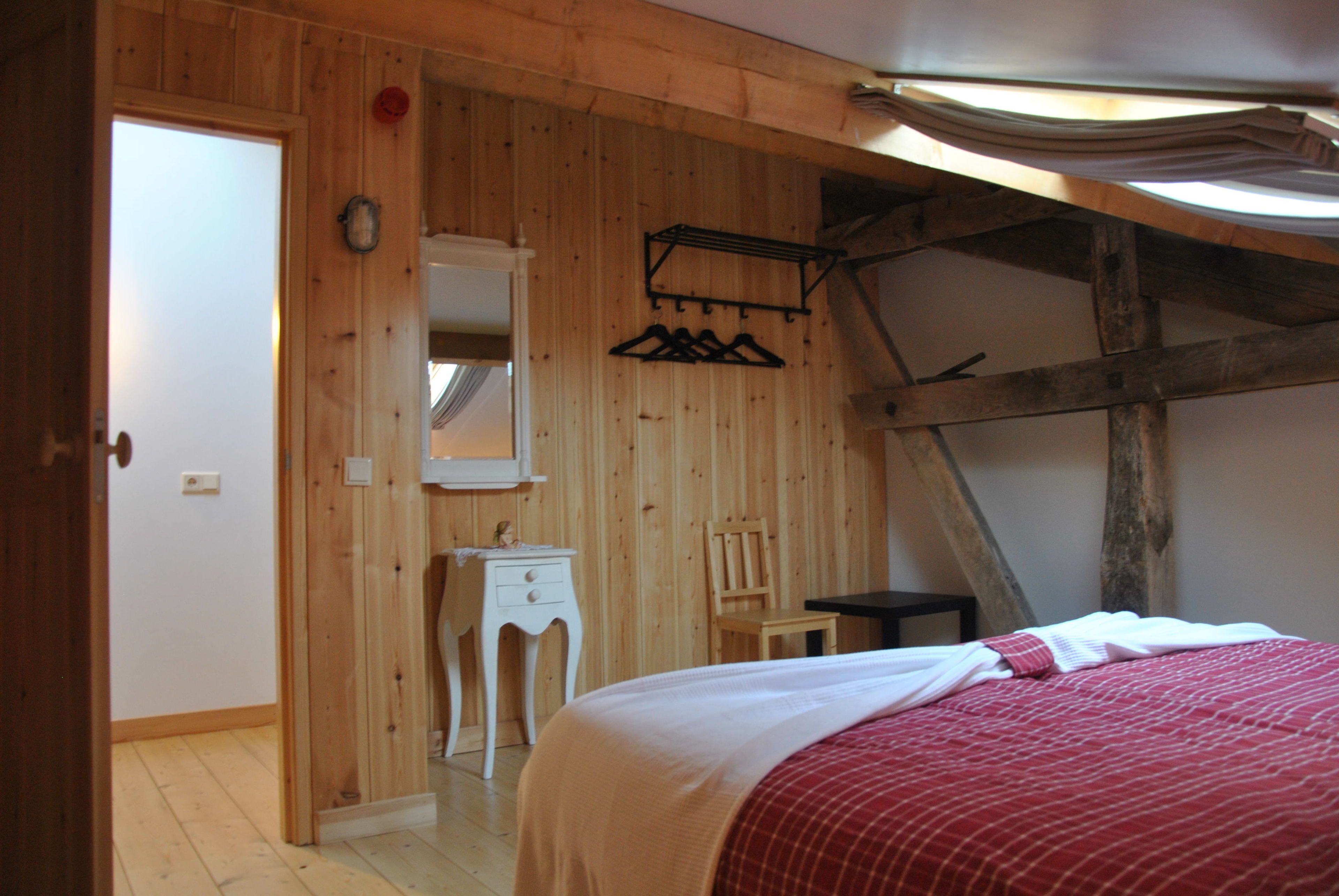 A bedroom with wooden walls, a bed with a red blanket, a wooden chair, a small table, and a mirror on the wall.