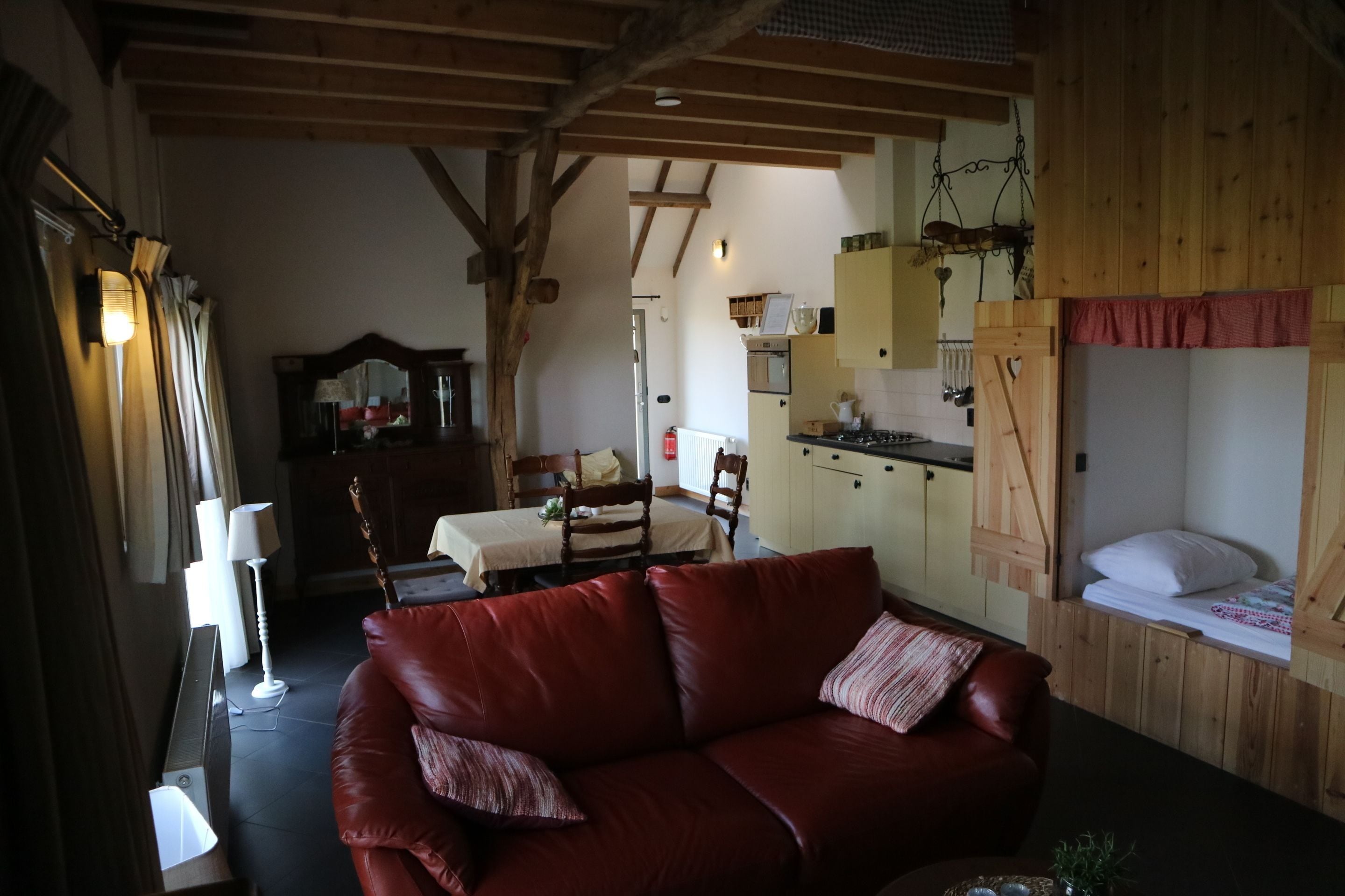 Una sala de estar acogedora con un sofá de cuero rojo, vigas de madera, una mesa de comedor con sillas y una zona de cocina con armarios de madera y un colgador para ollas.