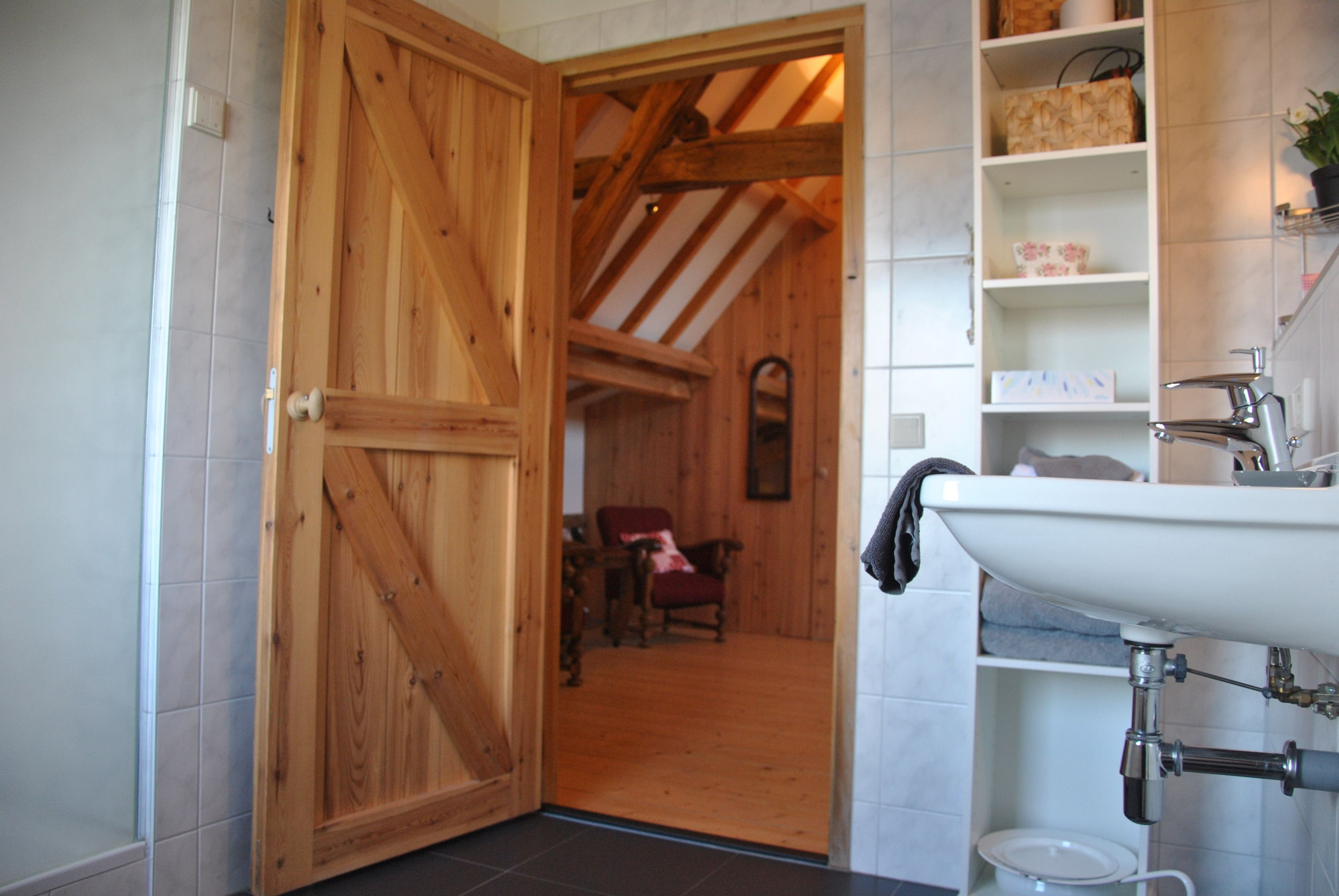 Een houten deur die leidt naar een kamer met een stoel en een plank met toiletartikelen.
