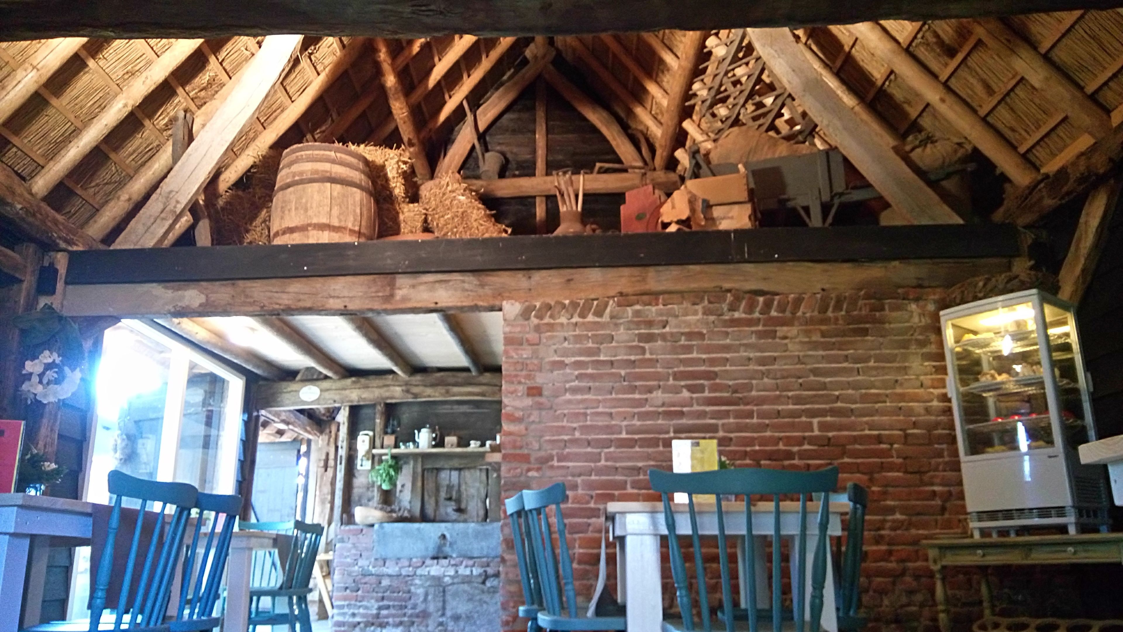 A rustic interior with exposed wooden beams, brick walls, and a brick fireplace. There are wooden chairs and a table in the foreground.