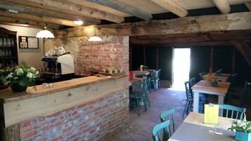 Un interior rústico con vigas de madera a la vista, pared de ladrillo y zona de bar con una cafetera. Mesas y sillas dispuestas para comer.