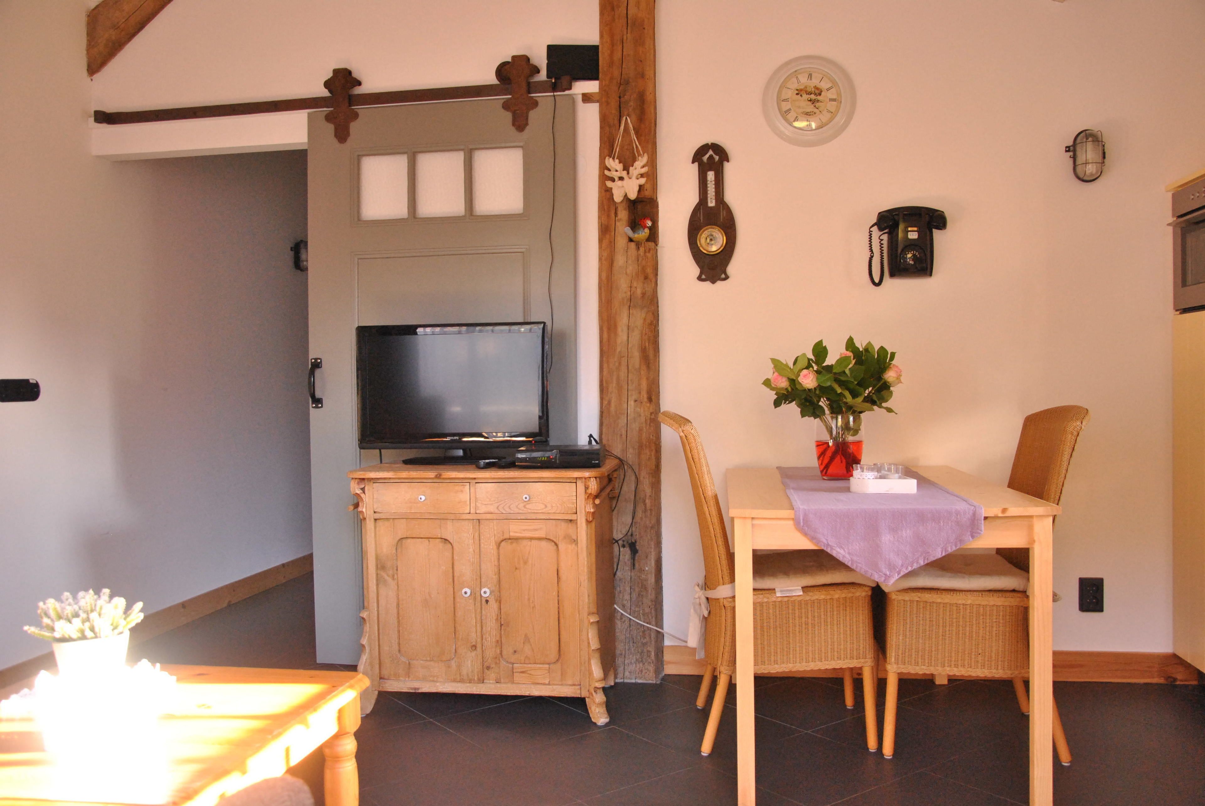 A dining area with a wooden table, wicker chairs, a television on a wooden cabinet, and a wall clock.