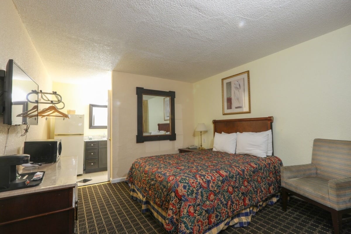 Una habitación de hotel con una cama, un escritorio con computadora, una silla, un espejo y un cuadro en la pared.