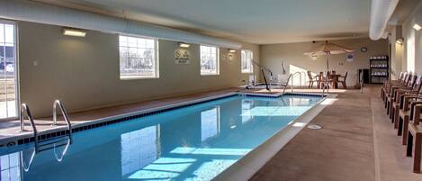 A large indoor swimming pool with clear windows, a lifeguard station, and a row of chairs along the poolside.
