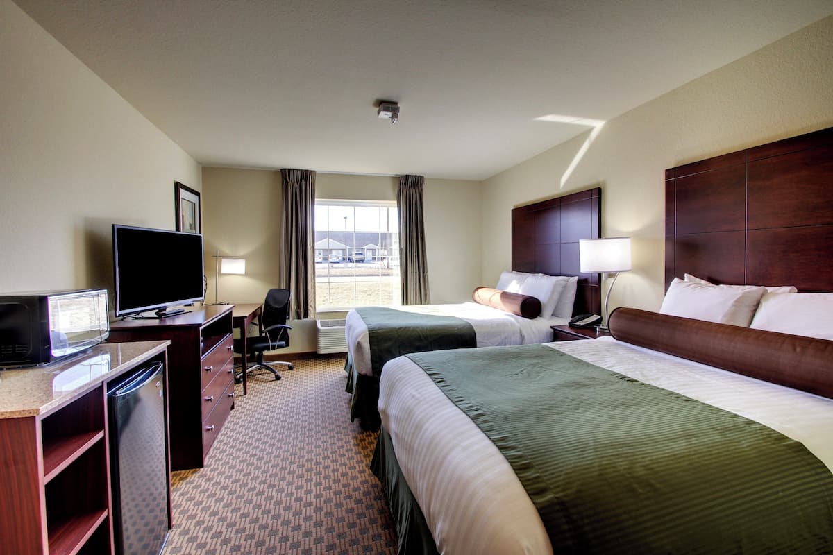 A hotel room with two beds, a desk with a TV, a refrigerator, and a view of the outside.
