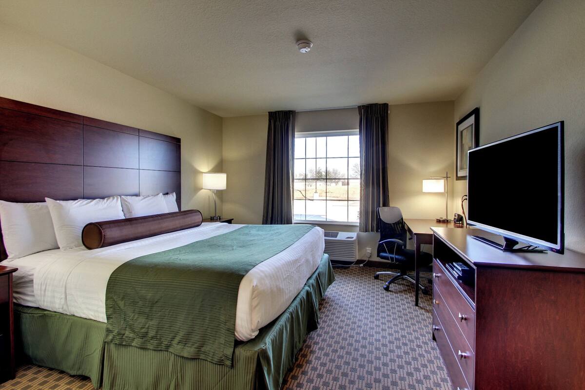 A hotel room with a large bed, a desk with a flat-screen TV, two bedside lamps, a window with curtains, and an air conditioning unit.