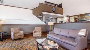 Un vestíbulo de hotel con una escalera, dos sillones estampados, un sofá gris y una mesa de centro de vidrio.