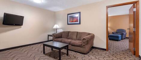 A room with a brown sofa, a small table, a TV mounted on the wall, a framed picture, and a patterned carpet.