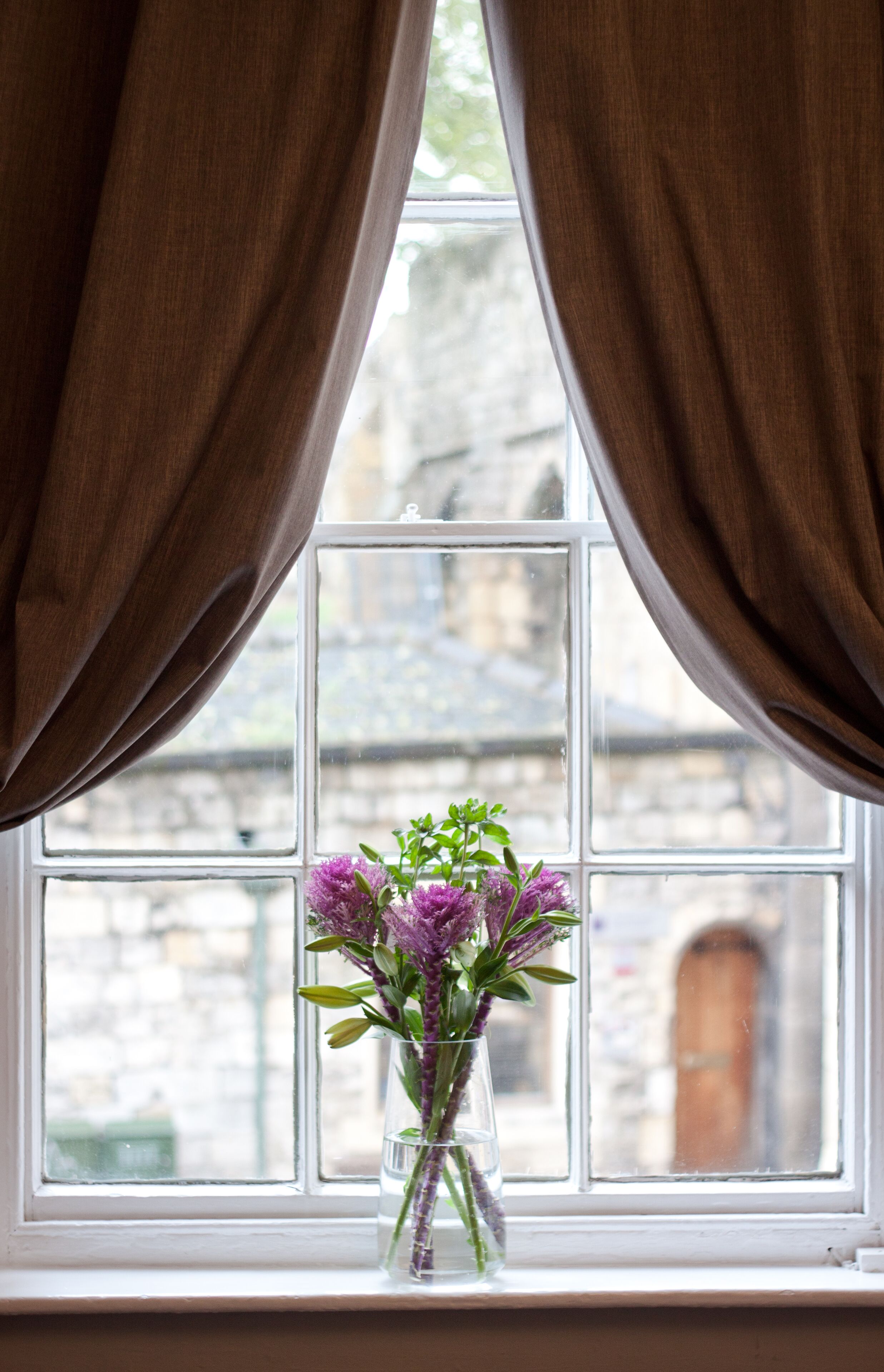 A window with brown curtains, a clear view of a stone building, and a vase with purple flowers on a windowsill.
