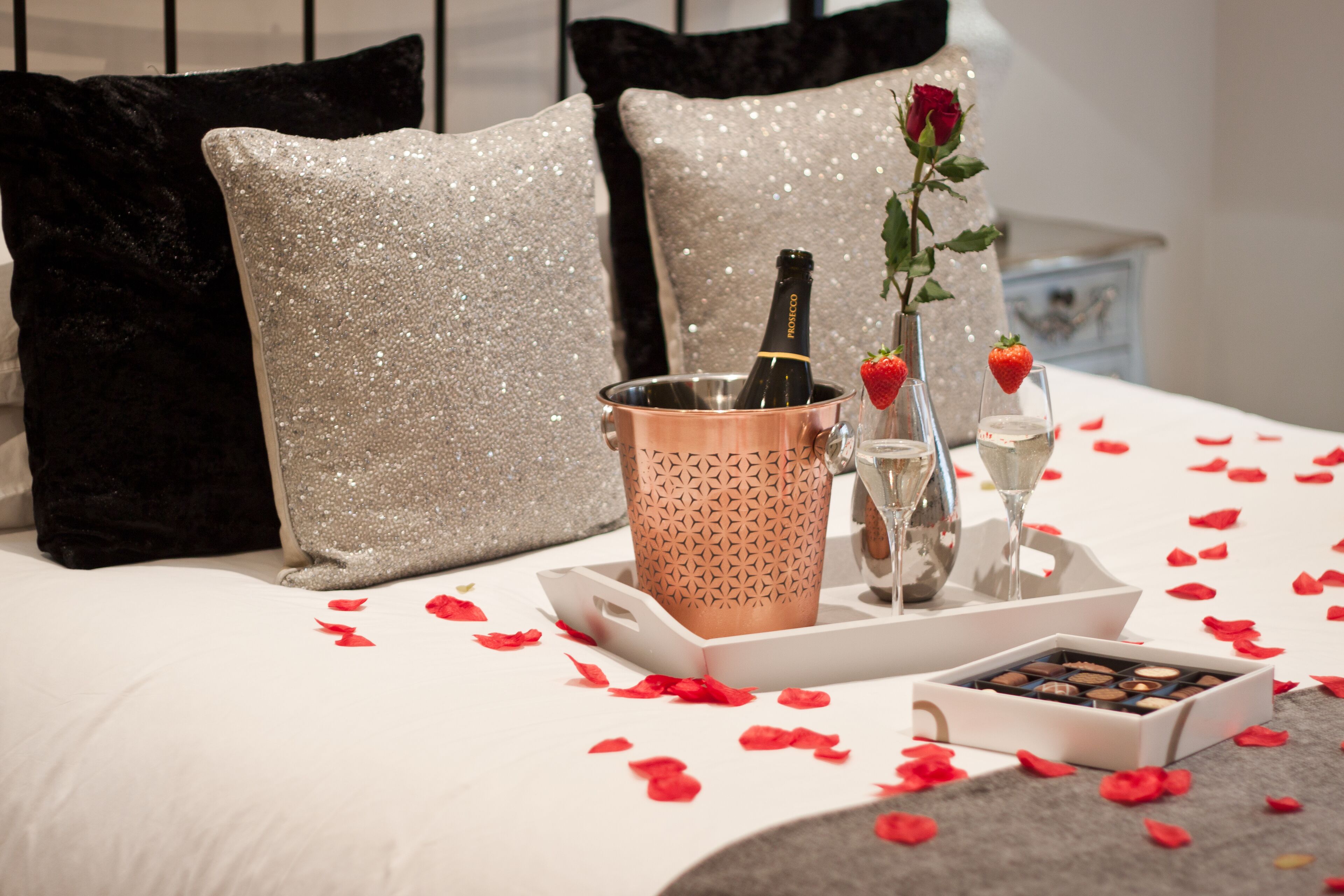 A bed with a champagne bottle, glasses, a rose, and chocolates on a tray.