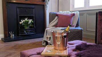 A living room with a fireplace, a flat-screen TV mounted above it, a plush purple sofa, and a wooden tray with champagne glasses.