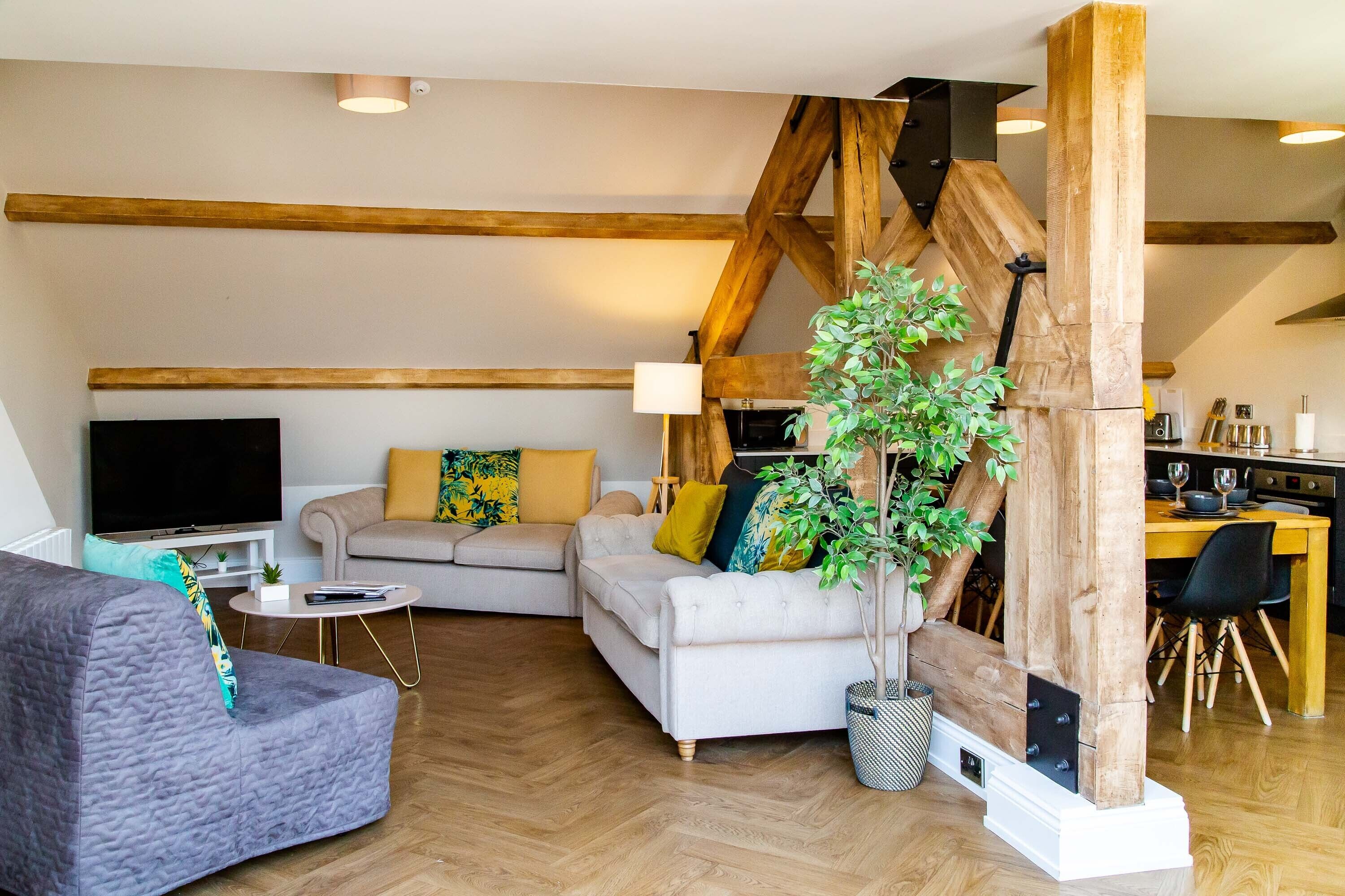 A living room with a sectional sofa, a TV, a dining area with a wooden table, and a large wooden beam structure.