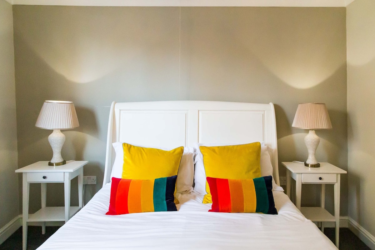 Un lit avec une literie blanche et des coussins colorés, flanqué de deux tables de chevet blanches sur lesquelles sont posées des lampes.