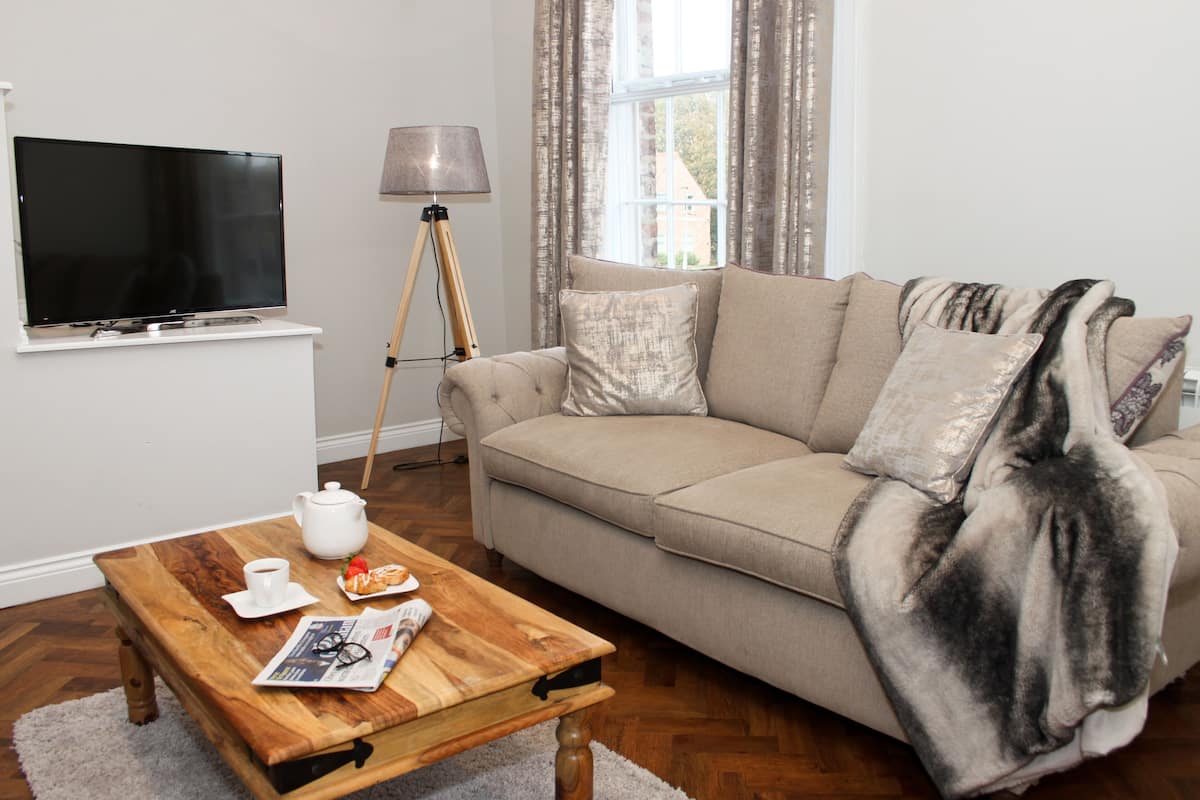 Un salon avec un canapé beige, une table basse en bois sur laquelle se trouvent une théière et une assiette de nourriture, un téléviseur fixé au mur et un lampadaire.