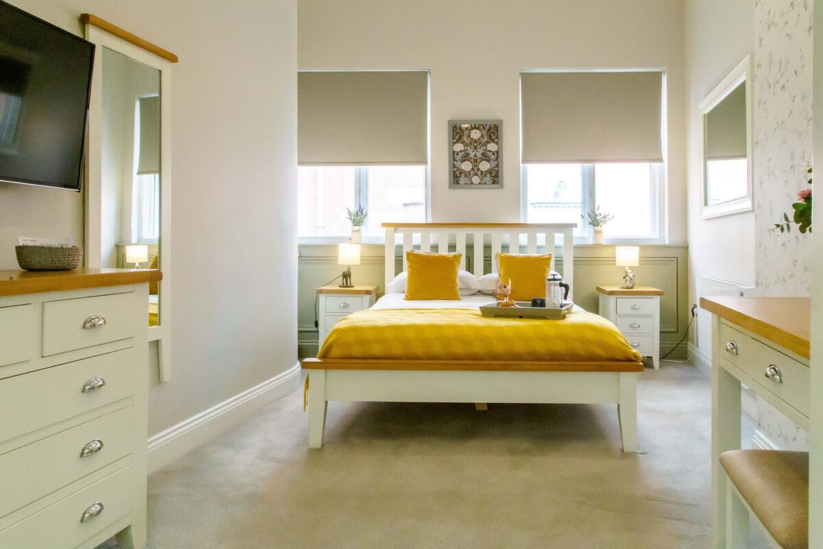 Une chambre à coucher comprenant un lit, des tables de chevet, une commode, un miroir et une fenêtre avec des stores.