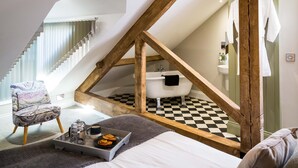 A bedroom with a sloped ceiling, a checkered floor, a bed, a chair, a tray with breakfast items, and a bathroom with a bathtub and a towel rack. - Goodramgate Apartments (York)