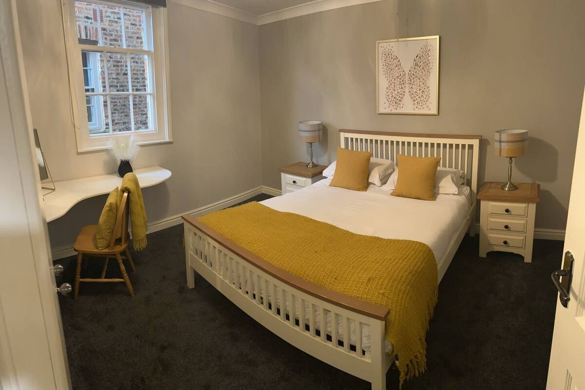 Une chambre à coucher comprenant un lit, des tables de chevet, une chaise, une fenêtre et une œuvre d’art accrochée au mur.