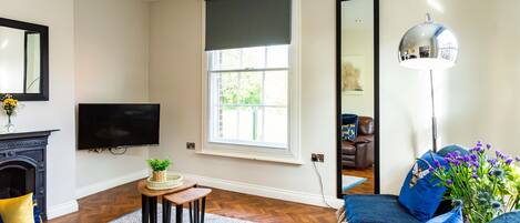 A living room with a fireplace, a flat-screen TV, a large window with a dark roller blind, a floor lamp, and a sofa with blue cushions.