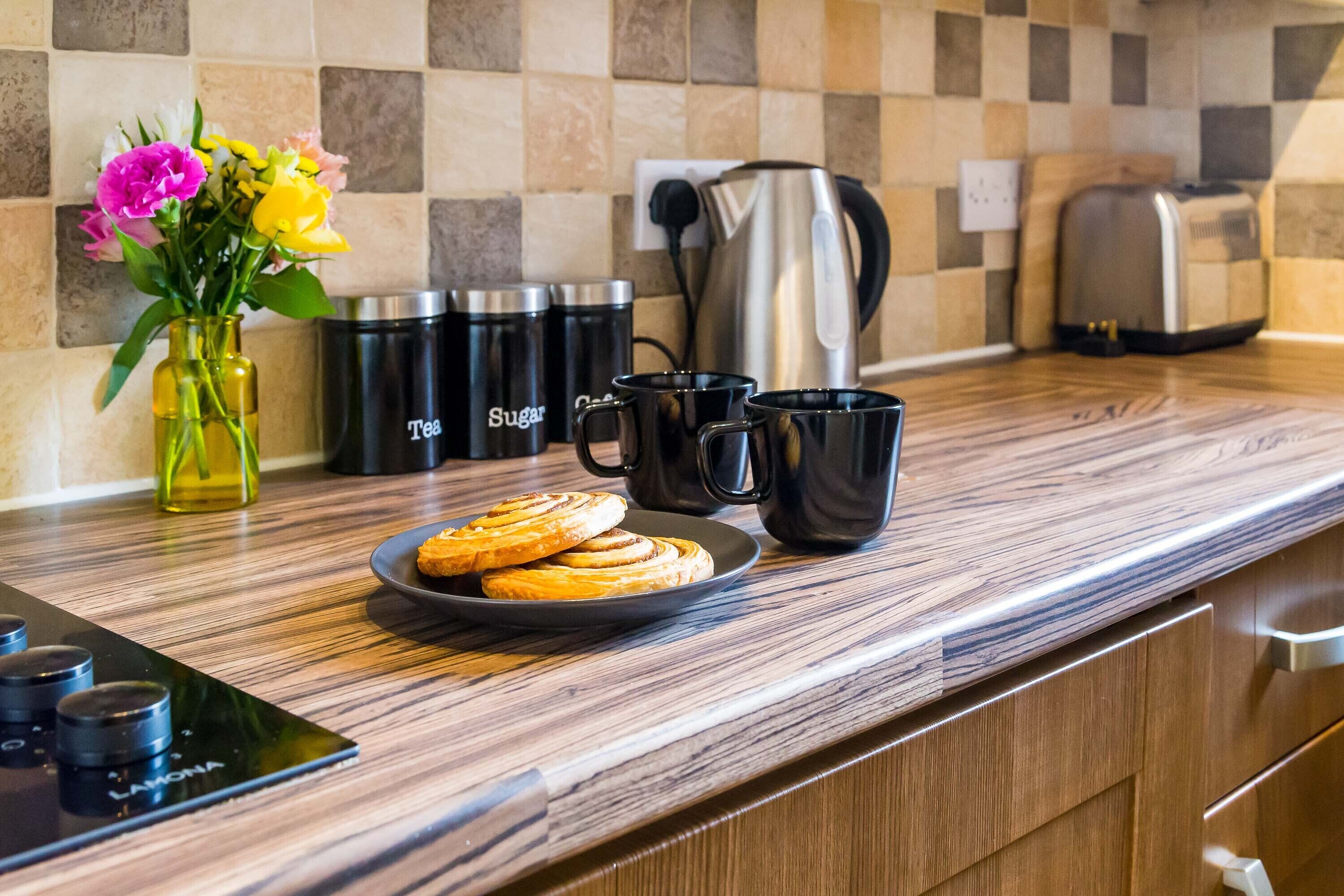 Una encimera de cocina con una cafetera, recipientes para té y azúcar, un plato de galletas y un florero con flores.