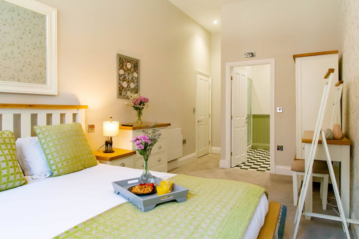 Une chambre d’hôtel avec un lit, une table de chevet avec une lampe, une petite table sur laquelle se trouve un plateau de petit-déjeuner, un tableau au mur et une porte qui donne sur une autre pièce.