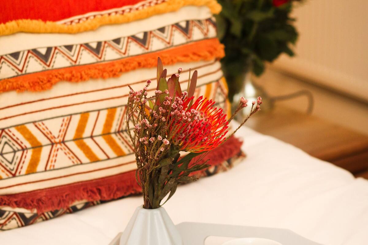 Un plateau avec un croissant, une fraise et une tasse de café, posé sur un lit agrémenté d’un oreiller à motifs et d’un vase de fleurs.