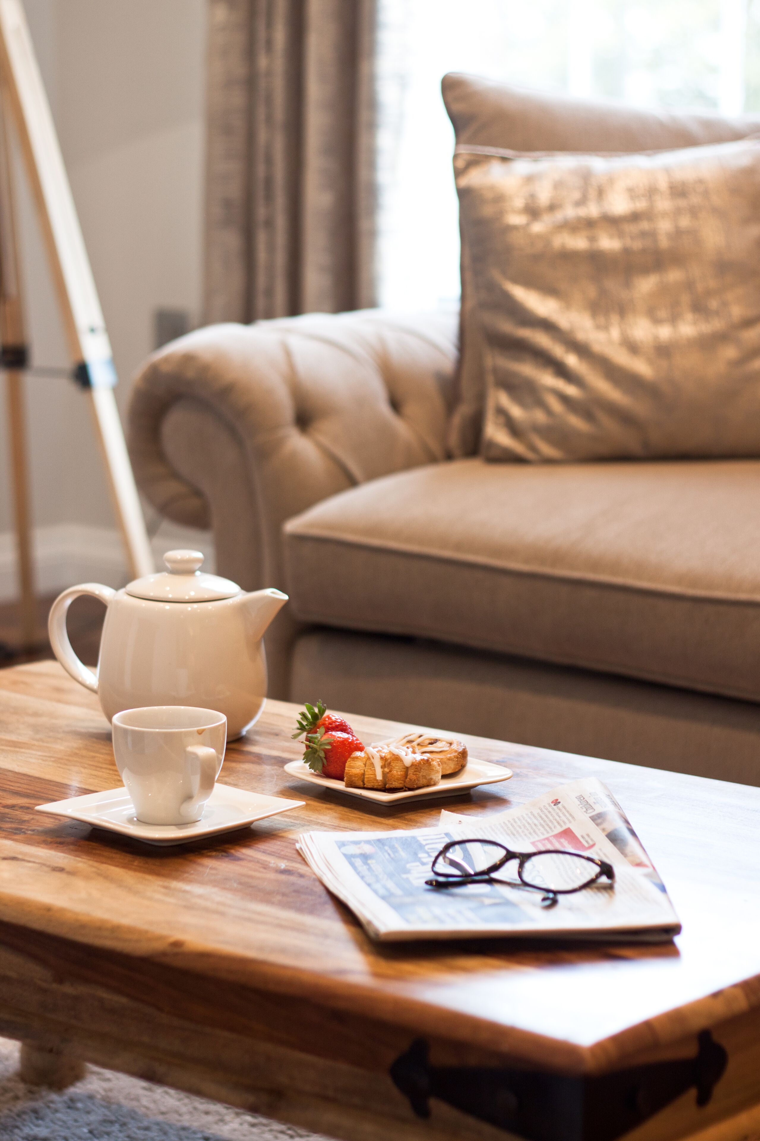 Una mesa de centro de madera con una tetera blanca, una taza, un plato con una fresa y un pastel, y un par de gafas sobre un periódico.