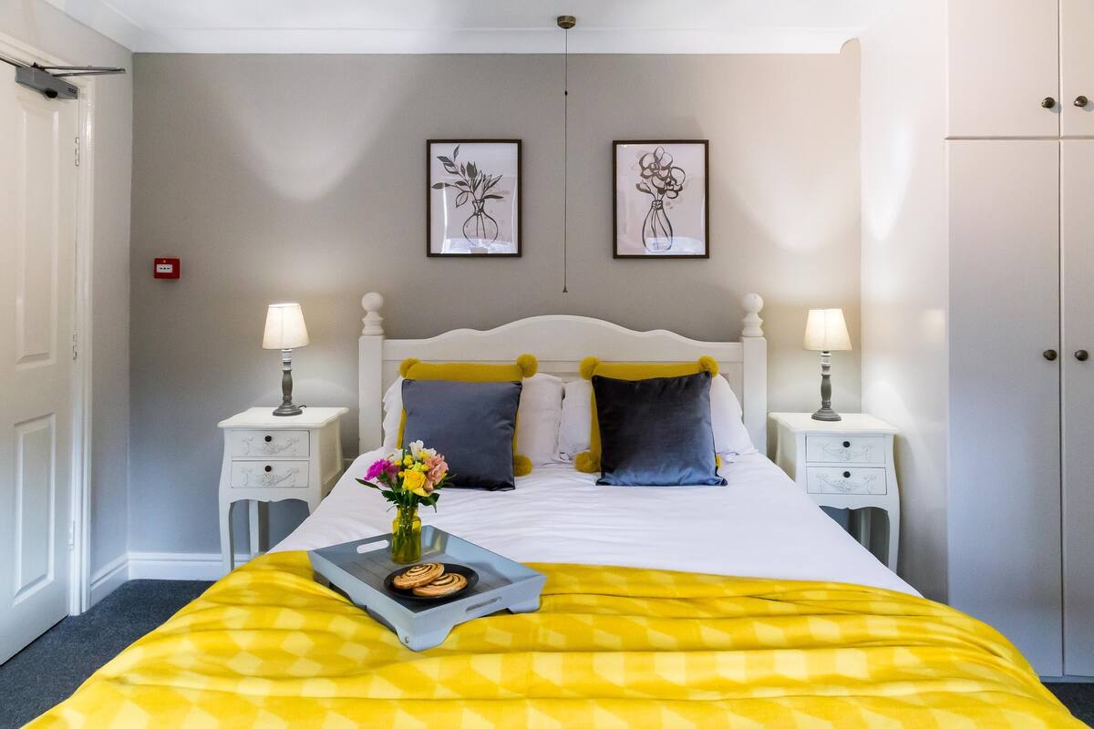 Une chambre d’hôtel bien rangée, avec un lit, deux tables de chevet, une petite table, une lampe, un plateau avec des biscuits et un vase rempli de fleurs.