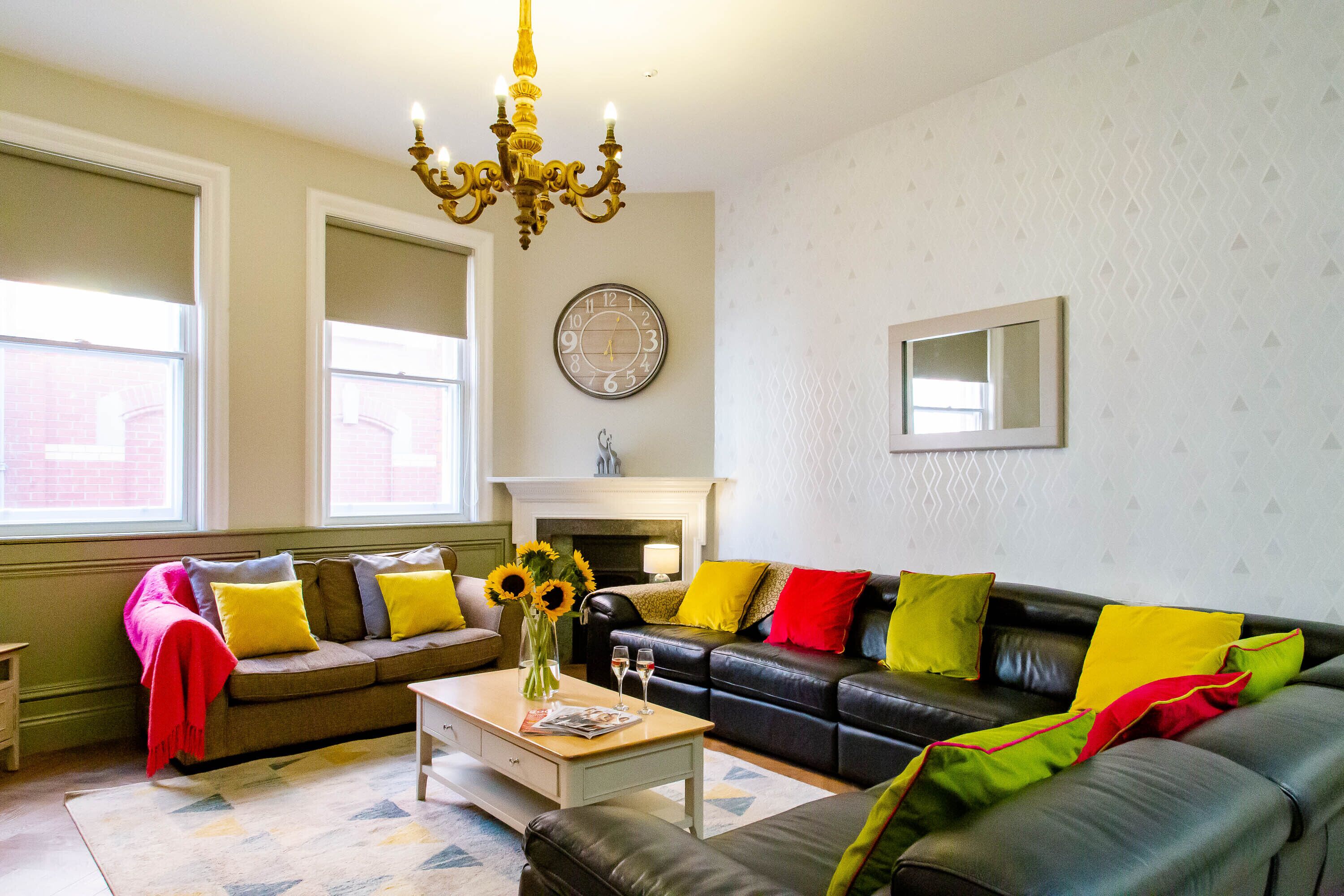 A living room with a fireplace, two sofas with colorful cushions, a coffee table with a vase and glasses, and a chandelier.