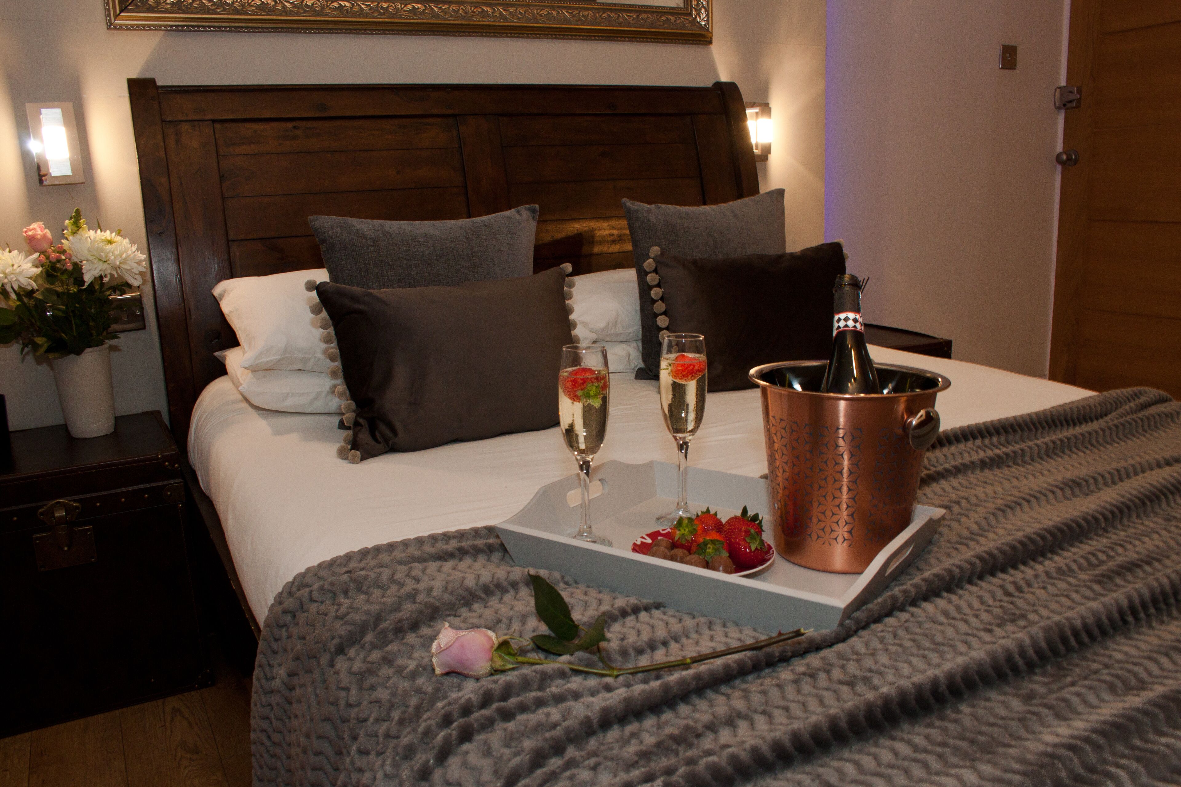 A neatly made bed with pillows, a bedside table with a vase of flowers, and a tray with champagne glasses and strawberries.