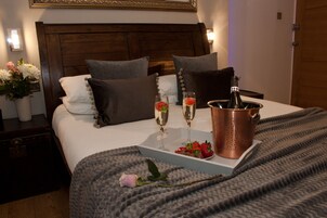 Una cama bien hecha con almohadas, una mesita de noche con un florero y una bandeja con copas de champán y fresas.
