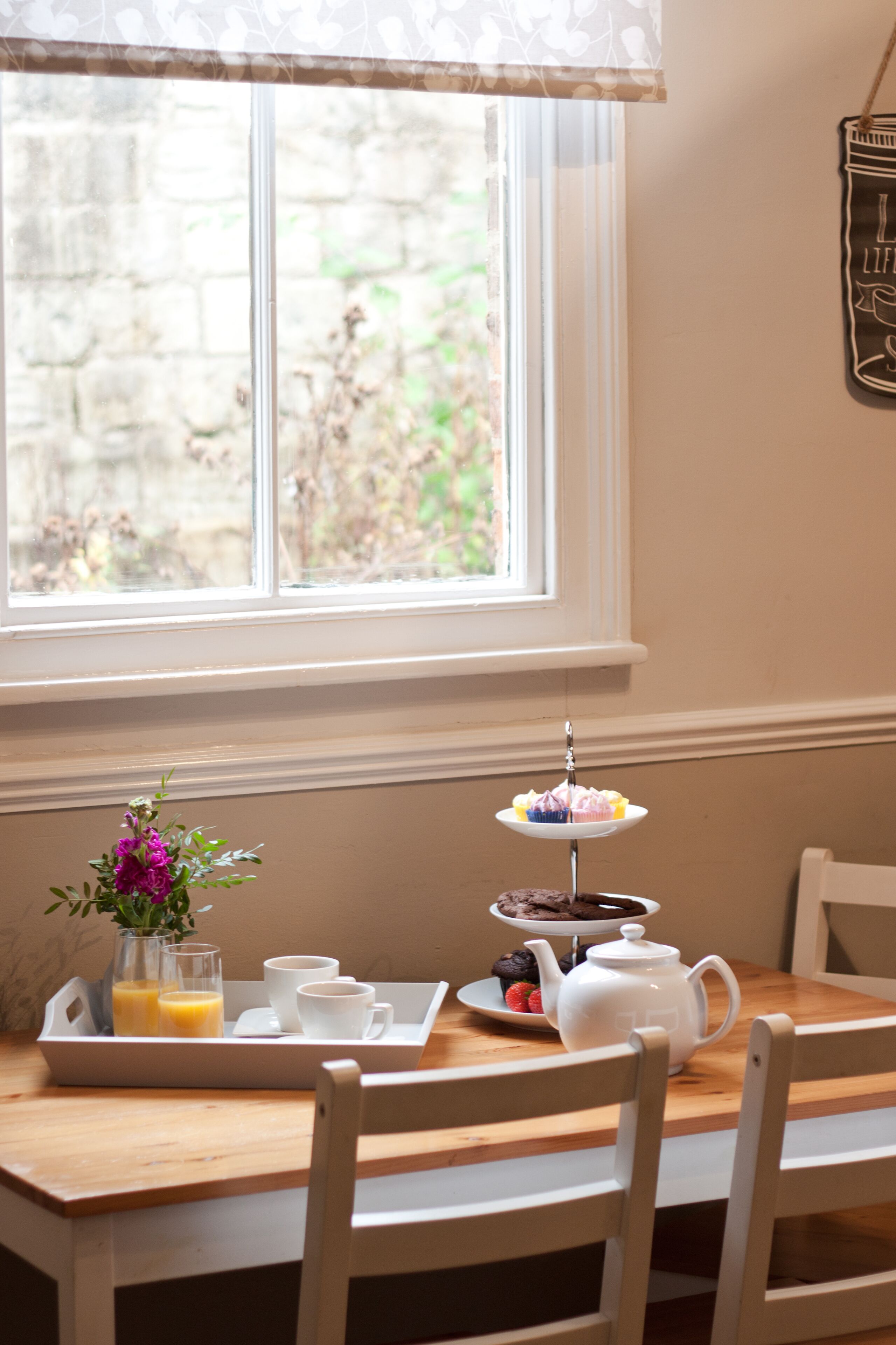 A breakfast tray with a teapot, cups, and a plate of food on a wooden table by a window.