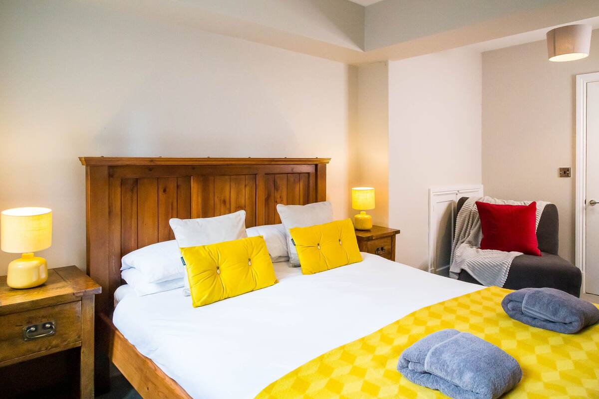 Un lit bien fait, avec une literie blanche et jaune, une tête de lit en bois et une couverture à carreaux. Il y a deux tables de chevet avec des lampes et un coussin rouge sur une chaise.