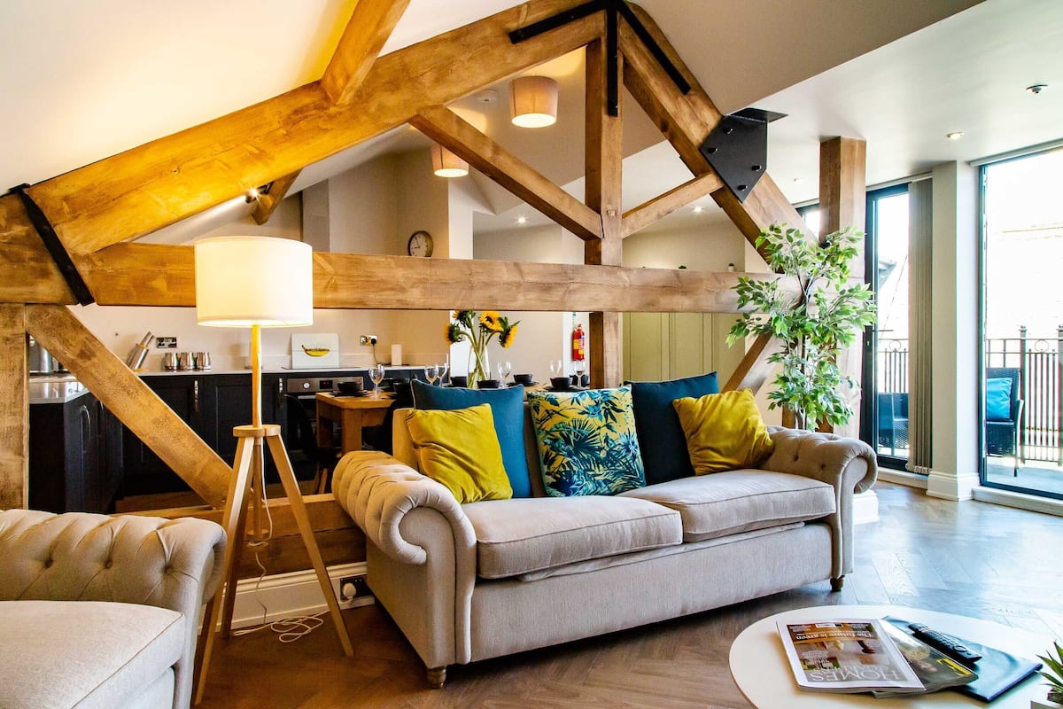 Un vaste salon agrémenté de poutres apparentes, un canapé avec des coussins aux couleurs vives, une lampe et, au fond, un coin repas.