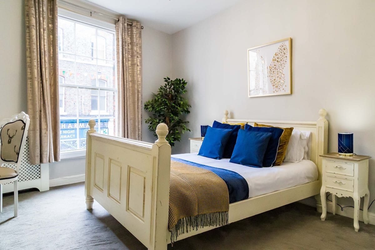 Une chambre à coucher avec un lit blanc, des coussins bleus et dorés, une table de chevet avec une lampe, et une fenêtre avec des rideaux.