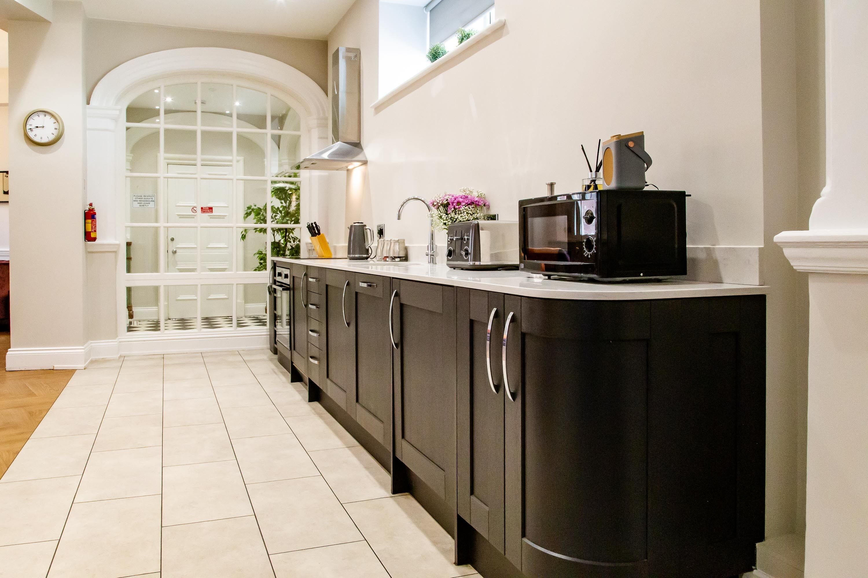 Een moderne keuken met donkere kastjes, een magnetron en een broodrooster. Er is een raam met uitzicht op een deur en een klok aan de muur.