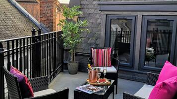 A rooftop terrace with wicker furniture, a table with a bottle and glasses, and a view of the cityscape.