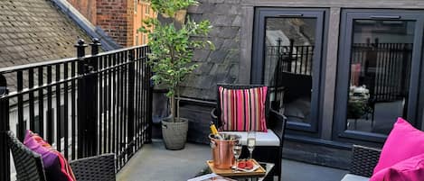 A rooftop terrace with wicker furniture, a table with a bottle and glasses, and a view of the cityscape.