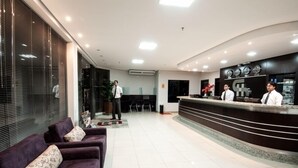 Un espace d’accueil d’hôtel comprenant un comptoir de réception, trois employés et un espace d’attente avec des canapés violets.